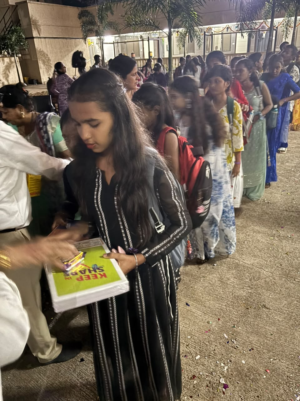 Children in a queue at an evening community event, receiving gift packets from a volunteer